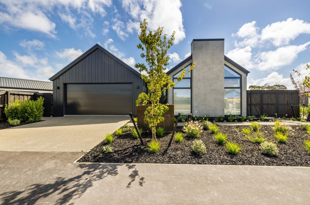 Oakbridge front exterior by Hallmark Homes Christchurch, displaying a contemporary architectural design with dark cedar vertical shiplap, light plaster panels, a gabled roofline, and a minimalist garden bed under a sunny blue sky.