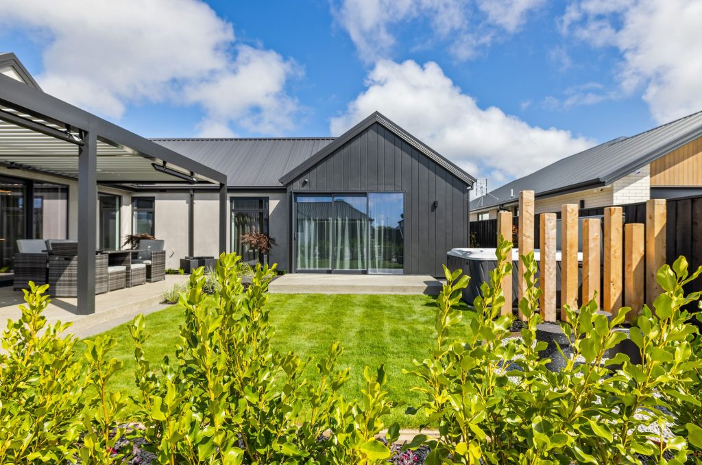 Back exterior of an architecturally designed Hallmark Homes build, with dark vertical cladding, plaster walls, a louvre pergola over an alfresco dining space, and feature timber screening.